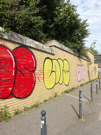 The image shows a brick wall covered in graffiti in Witten, Germany. The graffiti is in red, yellow, and pink bubble letters. The wall is located next to a sidewalk, which is paved with gray interlocking concrete pavers. There are several bollards along the sidewalk, and some vegetation is growing along the base of the wall. In the background, there are trees and a cloudy sky.