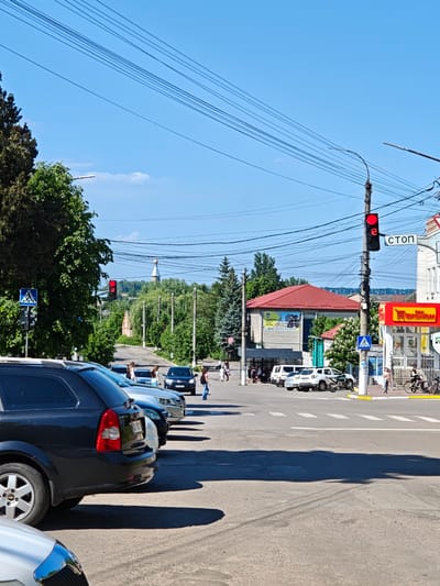 The image captures an urban scene in Illintsi, Ukraine. A red traffic light displaying "12" is mounted on a pole along with a "СТОП" (STOP) sign in Cyrillic. Several parked cars are lined up on the left side of the frame, with pedestrians using a crosswalk. A building with a red roof is partially visible on the right, adjacent to a storefront with a yellow and red sign, and a sign advertising Сипеди (bicycle). A smaller crosswalk is located in front of this shop. A church steeple can be seen in the distance behind the trees along the road. Overhead, many electrical wires crisscross the sky. The street appears to be in the intersection.