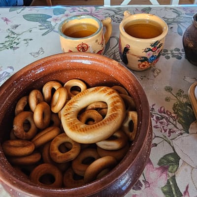 Two decorative ceramic mugs, one taller than the other, are placed on a tablecloth with a floral pattern. Both mugs are yellow with painted designs of flowers and a black chicken on the taller one, and a red circular motif on the shorter one. A large, rustic clay pot filled with small, ring-shaped baked goods, likely "baranki" or "sushki," is to the right of the mugs. The setting appears to be indoors, possibly a kitchen or dining area, with a glimpse of a wooden structure in the background suggesting a rural or traditional Russian home in Vybolovo, Russia. The lighting suggests daytime. The overall scene conveys a sense of a traditional, cozy atmosphere, perhaps preparing for a tea time or a simple meal. There is no visible text in the image.