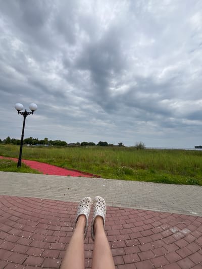 In Breytovo, Russia, a first-person perspective shot captures a person's legs, adorned with white patterned clogs, resting on a curved brick-paved area. The pathway is edged with red gravel and green grass. A dual-headed lamppost stands to the left, its white globes illuminated against a cloudy, overcast sky. Beyond the greenery, a body of water is visible, with a distant treeline and scattered buildings on the horizon.