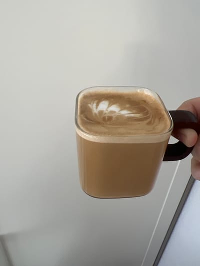 A close-up image displays a transparent glass mug containing a light brown coffee beverage, topped with white latte art forming a stylized leaf or fern pattern. The mug has a slightly rounded rectangular or square shape, with a dark brown or black C-shaped handle. A human right hand, featuring fair skin, grips the handle from the right side of the frame. The background consists of a plain, light grey or off-white wall occupying the upper and left portions. A sliver of a light-colored object, possibly an electronic device like a laptop, is visible in the lower right corner, partially obscured. The lighting is uniform and diffuse. The specific location within Kernen im Remstal, Germany, is not discernible from the visual information provided.
