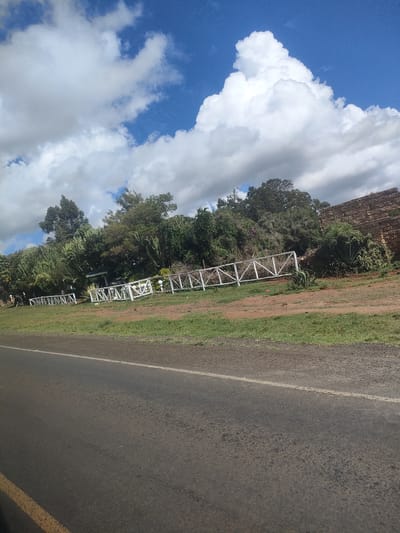 The image shows a section of a road in Kisima ward, Kenya under a partly cloudy sky. The road is asphalt with a faded white line marking. Adjacent to the road is a strip of grass, followed by a dirt area bordered by a white wooden fence. Behind the fence is dense vegetation, including trees and cacti. A section of a stone wall is visible in the background. No people or animals are visible in the immediate scene.