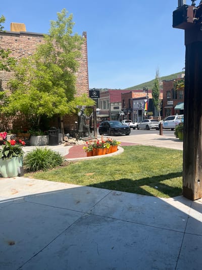 This image depicts a street scene in Park City, USA, bathed in the bright sunlight of a clear day. The perspective is from a covered walkway, looking out towards a street lined with brick buildings, indicative of a historic downtown area. A well-maintained grassy lawn borders a paved walkway, with planters containing colorful flowers adding visual interest.

In the background, several vehicles are parked along the street, including what appears to be a white police or service vehicle with "KONA BOURBON" written on it. Further down the street, a rainbow flag is visible, suggesting a diverse and inclusive community. The architecture of the buildings suggests a blend of old and new, with some featuring traditional brick facades and others having more modern storefronts. A black SUV is parked prominently in the foreground, with a statue and a bench visible near it.

The time of day is clearly daytime, with strong shadows cast by the overhead structure and trees. The overall atmosphere is serene and picturesque, suggesting a pleasant day for exploring the town. A sign with a circular logo and the word "AETHER" is also visible on one of the buildings. The scene is tranquil, with no visible human activity beyond the parked vehicles.