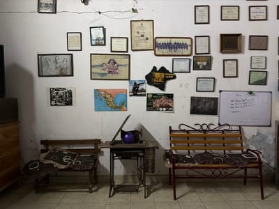 An interior scene depicts a tiled room with a collection of framed photographs and documents hanging on a white wall. A wooden chair with cushions sits to the left, while a vintage sewing machine rests on a small table. A metal bench with wooden slats and cushions is placed to the right. A whiteboard with handwritten notes is mounted near the bench. The room appears cluttered and worn, possibly a workspace or a storage area with personal memorabilia.