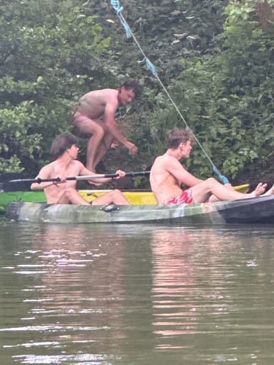 Three shirtless men are near water, possibly a lake. One man in red swim trunks is clinging to the side of a metal boat. Another man is inside the boat holding a paddle. A third man is seated on the bank, holding a blue rope connected to the boat. Green foliage lines the bank of the water. The water's surface shows reflection and some movement.