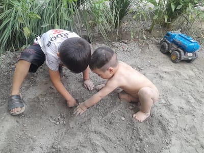 Two young children are playing in the dirt near a patch of greenery and a blue toy truck. The child on the left is older, wearing a white t-shirt with a red and black design, black shorts, and a grey sandal. The child on the right is younger and appears to be naked, with a shaved head. Both children are focused on the ground, with their hands digging into the dirt. The scene appears to be outdoors, possibly in a backyard or park, with natural lighting suggesting daytime. The overall mood is one of playful exploration. A blue toy truck is visible to the right of the children. The t-shirt of the older child has visible text that appears to say "RED STAR TEAM BRAND". The toy truck has the number "737" on its side and some license plate text that is unreadable. The location context is Danghara, Uzbekistan.