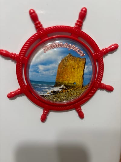 A red, plastic ship's wheel souvenir is displayed on a white surface. The wheel has eight spokes, each ending in a rounded knob. The center of the wheel contains a circular image, encased in clear plastic, depicting a rocky coastline with waves crashing against it. Above the image, in white cursive lettering, is the text "Дивноморское", which translates to "Divnomorskoe" in English. This text appears to be a place name. The image within the wheel shows a bright, sunny day with blue sky and white clouds. The overall scene suggests a seaside location and a souvenir from a vacation or trip. The lighting in the photo is soft, and there are no people or specific activities depicted. The setting is a plain white background, possibly a table or countertop, with a subtle shadow cast to the right, indicating overhead lighting. The souvenir is positioned slightly off-center, with the top of the wheel pointing towards the top of the frame.