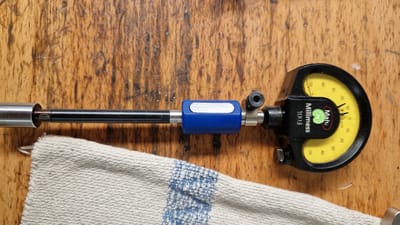 This media file displays a close-up, top-down view of a dial indicator and measuring probe resting on a wooden surface, likely a workbench. A piece of textured, off-white fabric with blue stitching is visible in the upper portion of the frame, partially covering the scene. The dial indicator, branded "Mah. Millimess 1003," has a yellow face with black markings indicating measurements in "μm" (micrometers). The needle on the dial points slightly past the "10" mark, towards the "20" mark, suggesting a measurement. Adjacent to the dial indicator is a measuring probe with a bright blue housing and a silver, elongated insert. The probe extends to the right, where it enters a silver cylindrical housing. The word "DIATEST" is visible on the probe's shaft. The wooden surface exhibits a worn, textured grain. The lighting suggests an indoor setting, possibly during daytime, with no specific weather cues. The overall impression is one of a precision measuring tool at rest, likely in a workshop environment in Ichenhausen, Germany. No people are visible.