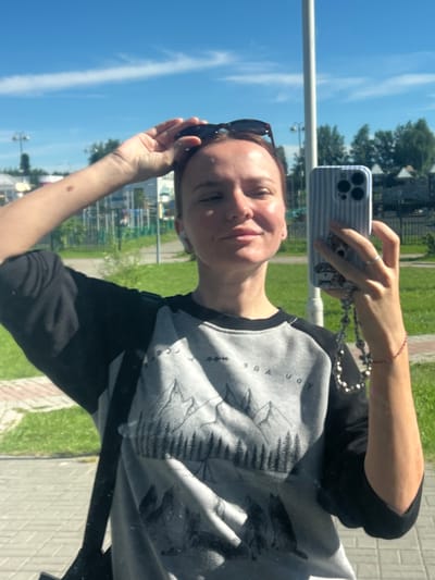 A woman is taking a mirror selfie outdoors on a sunny day. She is wearing a black and grey sweatshirt with a mountain and forest graphic. She has sunglasses on top of her head and is holding a smartphone in her right hand. The phone case is light blue with white stripes and a decal of a dog. She is also wearing a ring on her right ring finger and a red string bracelet on her right wrist. She has white wireless earbuds in her ears. Behind her, there is a green lawn, trees, buildings, and a blue sky with white clouds. A lamppost is visible in the background, and a paved walkway is in the foreground. The location is likely Mezhdurechenskiy, Russia, given the presence of typical Russian architecture and vegetation.