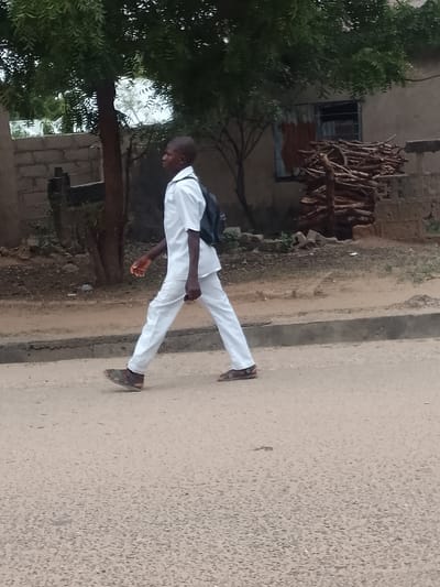 A young man wearing a white shirt, white pants, and sandals walks along a paved street in Biliri, Nigeria. He carries a black backpack. Behind him is a building with a window partially covered with corrugated metal, and a pile of wood. A tree stands to the left, and a brick wall is partially visible.