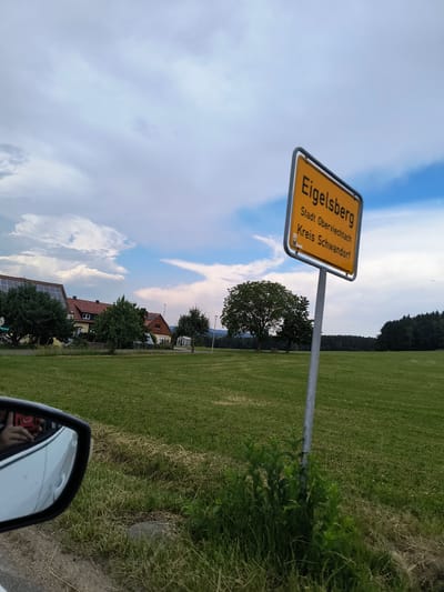 The image depicts a rural landscape with a prominent yellow sign that reads "Eigelsberg" with the text "Stadt Oberviechtach" and "Kreis Schwandorf" below it. This indicates the location is Eigelsberg, a part of the town of Oberviechtach in the Schwandorf district of Germany. The sign is mounted on a metal pole in a grassy field. In the foreground, a portion of a white car is visible, including its side mirror which reflects a person holding a red object, possibly a phone or camera. The background features a line of houses with red roofs and solar panels on one of the buildings. Further in the distance, there are trees and a dense forest. The sky is partly cloudy with a mix of grey and blue. The overall scene suggests a scenic drive through the Bavarian countryside.