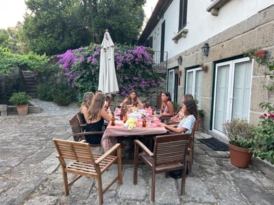 A group of seven young women are gathered around a table set for a meal on a stone patio. The table is covered with a colorful tablecloth and laden with bottles of beer, snacks, and food. The women are casually dressed and appear to be engaged in conversation and enjoying each other's company. Behind them, a white stucco house with dark shutters is visible, adorned with flowering purple bougainvillea. The scene is set in the city of Felgueiras, Portugal, likely at a private residence given the outdoor dining area and the proximity to the house. The overall atmosphere is one of relaxed outdoor dining and socializing in a rustic setting.