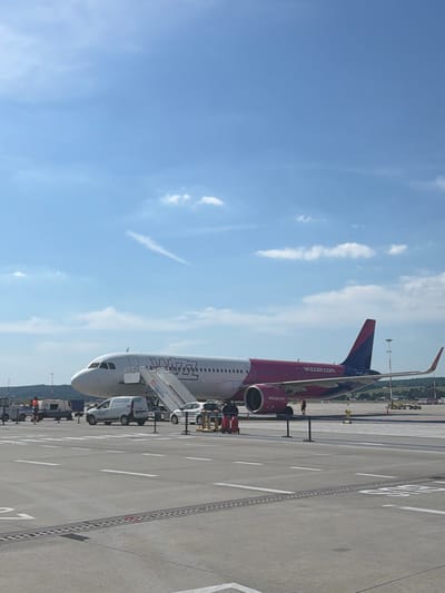 This media file shows a Wizz Air airplane parked on the tarmac at an airport. The airplane is predominantly white with a pink and purple livery featuring the Wizz Air logo and website address, "wizzair.com." A set of mobile stairs is attached to the front passenger door, indicating that passengers may be boarding or disembarking. A small white van with "LS SERVICES" written on its side is parked near the stairs.
The scene is set at an airport with a clear blue sky and scattered white clouds, suggesting fair weather. The sun is high, indicating it is likely daytime, possibly late morning or early afternoon. The ground is paved asphalt with white markings typical of an airport runway or taxiway. In the background, there are some trees and buildings, and what appear to be other airport vehicles and structures.
The orientation of the image is such that the airplane is viewed from the side, appearing to be standing upright in the frame. The perspective is from ground level, looking slightly upwards towards the airplane and the sky. There are no visible people engaged in any specific activity, though some individuals might be present on or around the vehicles. The overall atmosphere appears calm and functional, typical of an airport setting.
Visible text includes "WIZZ AIR" and "wizzair.com" on the airplane, and "LS SERVICES" and a number "309" on the van. There is also some text on the mobile stairs, partially obscured. The image is shot in Balice, Poland.