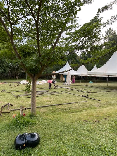 This photograph, taken in Makuyu, Kenya, captures an outdoor scene where preparations for an event are underway. The image is rotated, showing a landscape oriented vertically.
In the foreground to the left, a large tree with dense green foliage and a sturdy trunk dominates the frame. The tree's branches extend upwards, obscuring part of the sky. To the right, a black motorcycle helmet with the text "ITA" visible on its side rests on the vibrant green grass.
The mid-ground reveals the main activity: people are setting up multiple white canopy tents on a grassy field. Several sections of white tent fabric are visible, some already erected into structures, while others appear to be part of ongoing assembly. Underneath and around the tents, stacks of white plastic chairs are neatly arranged, indicating seating for an event.
Workers are actively involved in the setup. One individual, dressed in a pink shirt and dark trousers, is kneeling on the grass, seemingly engaged in assembling or arranging black metal poles that are scattered across the ground. These poles are likely components for the tent structures. Other figures are visible further back near the tents, also appearing to assist with the preparations.
The setting is an open field or garden with lush green grass. In the background, more trees and dense foliage line the perimeter, suggesting a natural and spacious environment. The sky above appears bright but overcast, indicating daytime with diffused light and no harsh shadows. The overall scene conveys a sense of industrious activity as infrastructure for an outdoor gathering is being established.