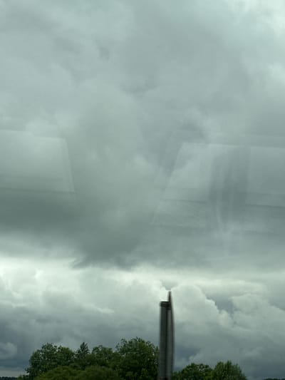 The image shows a cloudy sky over a line of trees. In the foreground, a dark, vertical pole, possibly part of a sign or structure, is visible on the right side of the frame. The clouds are heavy and gray, suggesting overcast weather. The trees below appear dense and green. The overall impression is one of a natural landscape under a somber sky. The location in the city of Issad, Russia, is not discernible from the visual content of the image.