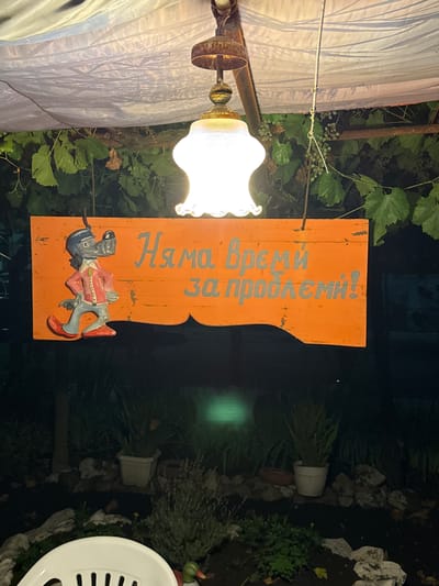 This is a nighttime outdoor photograph taken in Troyanovo, Bulgaria. In the foreground, a white plastic chair is partially visible. In the middle ground, an orange wooden sign with a cartoon wolf character and Cyrillic text is hanging from wires under a white tarp supported by a rusty metal frame. The sign reads "Няма време за проблеми!" which translates to "No time for problems!". Behind the sign, a garden area is visible with various plants in pots, rocks, and ground cover. A bright light hangs above the sign, illuminating the scene. The background is dark, suggesting it is nighttime.