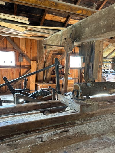 The image shows the interior of an old-fashioned saw mill, likely located in South Dundas, Canada. The structure is made of rough-hewn wood, with visible beams supporting a sloped roof covered in wooden planks. Light streams in from two windows, illuminating the dusty, sawdust-covered floor and machinery.

The primary focus is a large, mechanical saw mill with a log positioned on a track, ready to be cut. A circular saw blade is positioned above the log, held by a heavy metal frame. Gears and chains are visible, indicating the power mechanism of the saw. Various wooden planks and lumber are stacked around the mill, some on the floor and others along the walls.

In the background, more wooden shelves are visible, holding what appear to be tools and equipment related to the lumber industry. The overall impression is one of a working historical site, preserved to showcase the processes of early lumber production. There are no people visible in the image.