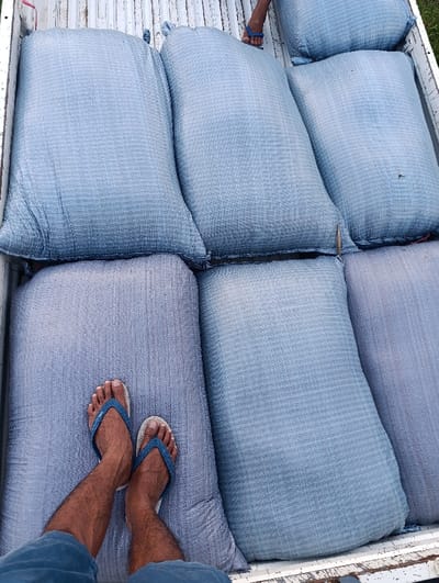 The image shows a truck bed filled with large blue and gray sacks. The sacks are stacked in two rows of three, with the top row slightly offset from the bottom.  A person's legs are visible in the lower left corner of the image, with the right foot resting on one of the sacks and the left foot wearing a blue flip-flop. The scene is likely in a rural area near Arjasa, Indonesia, as the coordinates provided (latitude: -7.775, longitude: 114.134) place it in the middle of the island of Java. The surrounding environment is not visible in the image.