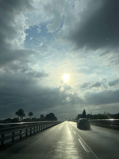 The image captures a view from a vehicle traveling on a wet highway, likely in or near Barletta, Italy, given the context. The sun is shining brightly through a gap in the dramatic, dark clouds, casting a strong reflection on the wet asphalt. The road ahead curves slightly to the right, with a guardrail lining the left side. A dark-colored car is visible in the right lane, ahead of the viewer's vehicle. Palm trees and other foliage are silhouetted against the sky on the right side of the road, with a few buildings and a church spire visible in the distance on the horizon. The overall atmosphere suggests a dramatic transition in weather, possibly after a rain shower.