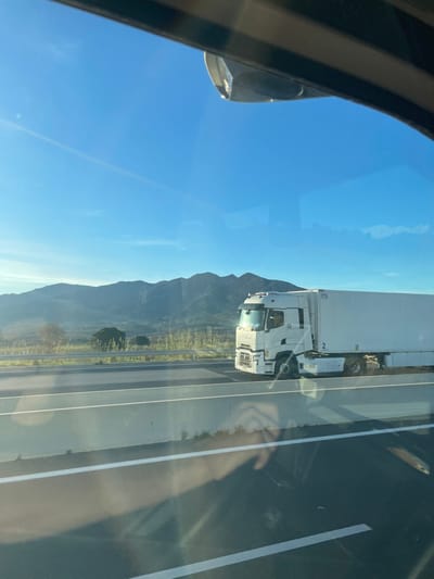 The image depicts a white Renault T-range semi-trailer truck traveling on a multi-lane highway, viewed from inside another moving vehicle. The truck's cab bears the name "Felix," and the white box trailer has the number "2" marked on its side. In the background, a range of dark green mountains rises under a clear, bright blue sky with faint atmospheric hazing and a subtle contrail visible high above. Fields with sparse vegetation and a few trees are situated between the highway and the mountains. The highway features at least two lanes of dark asphalt with white lane markings and a concrete median barrier. Reflections are visible on the window through which the photograph was taken, including a human hand holding a device, presumably the camera, and parts of the vehicle's interior. A portion of a side-view mirror is visible in the upper frame. The scene is located in Tresserre, France.