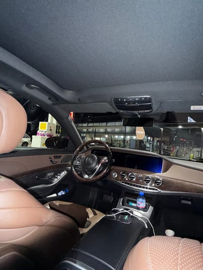 The image shows the interior of a luxury car, likely a Mercedes-Benz S-Class, parked at a gas station. The interior features brown leather seats and dark wood trim. The dashboard includes a large infotainment screen, climate control vents, and a steering wheel with the Mercedes-Benz logo. A bottle of water and a pair of glasses are visible on the center console. Outside the car, a gas pump and a store can be seen. The scene is lit by artificial lights, suggesting it is nighttime. The specific location of the gas station in Grevena, Greece, is not discernible from the image itself.