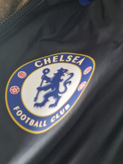 The image shows a close-up of a navy blue garment with the Chelsea Football Club crest prominently displayed. The crest features a white background with a blue lion holding a staff, surrounded by the words "CHELSEA FOOTBALL CLUB" in blue lettering on a yellow border. The garment appears to be made of a synthetic fabric and has a zipper visible along the edge. There are no people visible in the image. The location is not specified but the badge suggests Pity Me in the United Kingdom would be a plausible location.