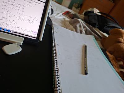 This is a close-up shot of a study area. Dominating the scene is a notebook with spiral binding, partially open to pages filled with handwritten mathematical equations. A 2H pencil rests on the left-hand page. To the right of the notebook lies a tablet, displaying a similar set of mathematical problems and equations. An eraser is placed near the tablet. Part of a stuffed animal is visible in the lower left of the image. Below the notebook is a messy area with a bag and some other items. The scene appears to be in a personal study space, possibly at home. There is no information to suggest that this image was taken in Voiniagovo, Bulgaria.