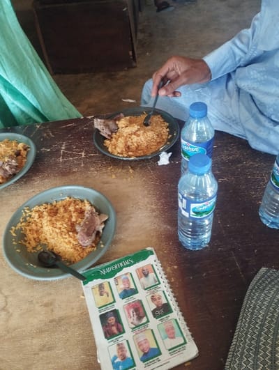 The image shows a wooden table with four plates of food, two bottles of water, and a green booklet. The food is rice and meat. A person is sitting at the table, wearing a blue robe and holding a spoon, taking a bite of food. The person’s right leg is visible, and their left leg is off-screen. An open book with a green cover, titled "LEAD OFFICIALS", lies on the table. The book contains photos of people. The table is set in a room that appears to be a restaurant. The scene is captured from a slightly elevated angle. The location is Kaltungo, Nigeria (latitude: 9.816, longitude: 11.314).