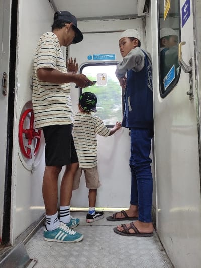 The image captures three individuals standing inside a train carriage during daylight hours. The interior of the train is predominantly white, with a metallic, textured floor and large windows revealing a glimpse of green foliage outside.
In the foreground on the left, an adult male stands, wearing a black baseball cap backwards, a short-sleeved striped t-shirt (white, light brown, and black stripes), black shorts, white socks with black stripes, and light blue Adidas sneakers. He is holding a smartphone in his right hand and appears to be looking down at it, engaged with its content. His left hand is slightly raised. Near his feet, a red circular object resembling a handwheel or valve is visible on the lower wall.
To the right of the adult, a younger child is positioned, mostly with his back to the camera. He wears a striped t-shirt similar in pattern to the adult's, light-colored shorts, black socks with white accents, and black sneakers with a white checkerboard-like pattern. He wears a black baseball cap with some stylized text on it. His arms are bent, and his hands are held up.
Further back, in the center of the frame, an older child stands facing a window, looking outwards. He is wearing a light grey hooded top over a dark blue jacket or shirt, dark blue long pants, and brown sandals. A white skullcap (peci) is on his head. His reflection is visible in the window above him.
Visible textual elements include "ADIDAS" on the adult's sneakers and "KECAMATAN BEJI" on a sticker affixed to the window. Other icons, such as a blue square depicting a person (often signifying priority seating) and a yellow charging symbol, are also present on the window stickers. The scene conveys people traveling by train, with individuals engaged in personal activities like using a phone or observing the view. The overall mood appears calm and neutral.