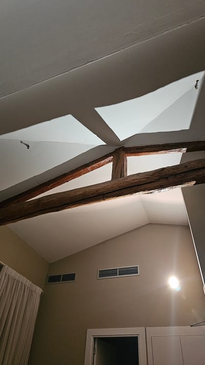 This is a low-angle, interior shot of a room with a vaulted ceiling. Two large, rough-hewn wooden beams cross diagonally across the upper portion of the image, supported by a central wooden post. Above the beams, the white ceiling is illuminated by geometric light patterns. On the tan-colored walls below, two rectangular air vents are visible, along with a bright spotlight. A beige curtain hangs on the left side of the image, and a doorway with a white frame is visible at the bottom. The scene suggests an architectural interior, possibly in a building with exposed beams and a modern ventilation system. The location is not specified in the image itself.