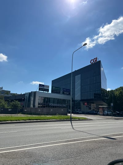 The image captures a sunny daytime view of a modern commercial building with a distinctive dark glass facade and a red "CGI" logo at its apex. The building is situated in the city of Tartu, Estonia. The foreground features a two-lane asphalt road with white lane markings, and a sidewalk with a grassy strip and fencing. Several cars are visible on the road and in front of the building. Signs on the building indicate various businesses, including "PORANDASALONG," "WISO," "evecon," "RM STUDIO," "Keskerakond," and "thektor light." A street lamp with a long shadow stretches across the road. The sky is bright blue with scattered white clouds and the sun is high, causing glare. Trees line the background behind the building. A bus stop shelter is also present on the right side of the image.