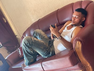 In an interior setting in Ogbunu, Nigeria, a single male subject is observed reclining on a reddish-brown, multi-seater upholstered sofa. The subject is dressed in a white tank top and distressed blue jeans, appearing barefoot. A square-faced wristwatch with a dark band is present on his left wrist. He holds a dark-colored mobile phone with both hands, directing his gaze downward towards the device's screen. The background consists of a textured, light-colored wall. A dark textile, consistent with a curtain, and a dark blue object are partially visible on the left side of the frame.