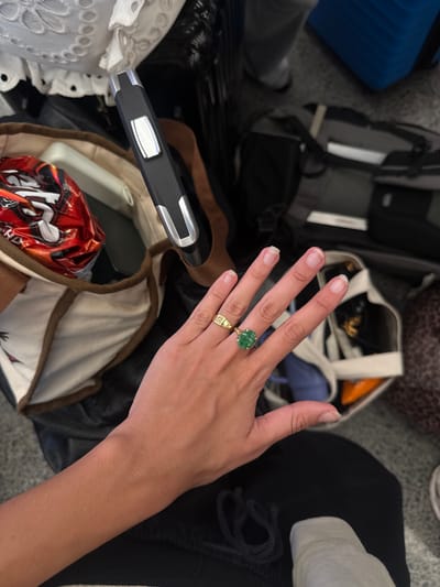 The image depicts a person's hand adorned with two rings against a backdrop of luggage and travel-related items. The hand, with manicured fingernails, features a ring with a large green stone on the ring finger and a gold signet ring on the pinky finger.

The background consists of various items, including luggage, bags, clothing, and what appears to be a package of Skittles candy. There is a suitcase handle extending across the bottom of the photo.

There is insufficient information to determine the location, as the items in the frame do not contain any identifying information relating to Honolulu or the United States.