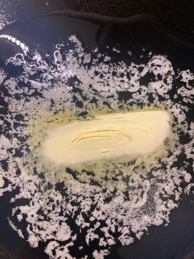 A close-up, overhead shot shows a stick of butter melting in a dark, possibly non-stick, pan. The butter is a pale yellow and is beginning to liquefy around the edges, forming a pool of melted fat. The surface of the pan is covered in a dark, oily substance with white, foamy-looking patches and small bubbles, suggesting the butter is beginning to brown or that other ingredients have been added. The lighting appears to be artificial, creating bright highlights on the wet surfaces. The scene is set within a kitchen environment in Lütjenwestedt, Germany.