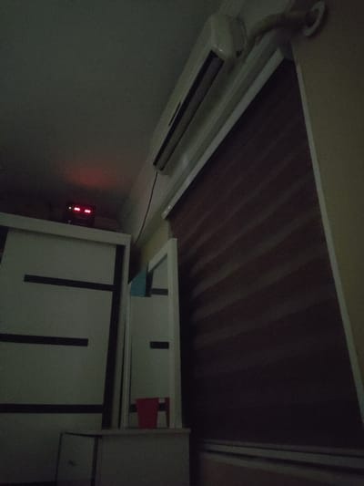 The image depicts an indoor scene. A white air conditioner unit is mounted high on a wall, partially visible. To the left, there is a white wardrobe with dark horizontal stripes. A digital clock with red LED numerals is positioned on top of the wardrobe, displaying "00:00". To the right of the wardrobe, a mirror or a white cabinet with a reflective surface is present, with a glimpse of a red cup on a shelf. The background features a dark, horizontally striped blind or curtain covering a window. The lighting is dim. The image does not provide any information to identify its location within the city of Кипчок, Tajikistan.