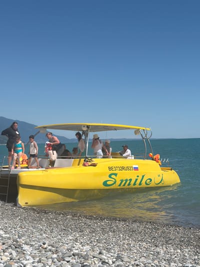 The image depicts a bright, sunny day in Amzara, Abkhazia, with a yellow catamaran boat docked on a pebble beach. The boat is branded with the word "Smile" in green lettering and a smiley face icon. Several people, including adults and children, are boarding or standing on the boat. One woman in a black top and a child in a turquoise swimsuit are at the front of the boat, while other individuals are visible on the deck and near the entrance. The sea is a calm, deep blue, and mountains form a backdrop against the clear sky. The scene suggests a recreational outing or a boat tour.