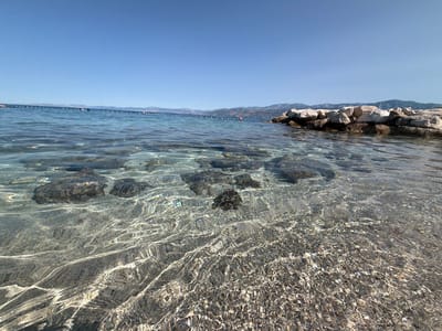 The image depicts a shallow, clear body of water with a rocky seabed visible beneath the surface. The water is calm, with gentle ripples reflecting the sunlight. In the background, a breakwater made of large stones extends into the water. Beyond the breakwater, a coastline with hills and mountains is visible under a clear blue sky. A line of buoys stretches across the water in the distance. The scene appears to be a beach or coastal area. The location is likely Općina Postira, Croatia, given the characteristic coastal landscape and clear Adriatic Sea.
