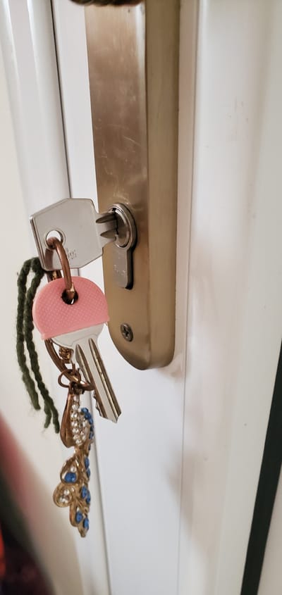 A close-up shot shows a set of keys inserted into a door lock. The keys are attached to a keychain featuring a decorative butterfly charm adorned with clear and blue rhinestones. One key has a pink, textured cover, and another key is partially visible, suggesting multiple keys are on the ring. The lock mechanism is mounted on a metallic plate that appears to be part of a door. The background consists of a white, slightly textured surface, possibly the door itself or a wall. The lighting suggests an indoor setting. The overall impression is one of everyday use, with the decorative keychain adding a personal touch to the functional object. There is no visible text in the image. The provided location context of Νέα Σάντα, Greece, does not offer any direct visual cues within the image itself.