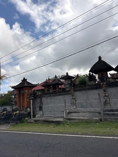The image shows a street-side view of a building with traditional Balinese architecture in Susut, Indonesia. A grey concrete wall with stone accents and decorative pillars runs along the property line. Several structures with thatched roofs and ornate detailing are visible behind the wall, including a prominent one with a pointed roof and a small bell. To the left, a red-orange gated entrance is flanked by two dark statues with gold trim, resembling lions. The ground in front of the wall is covered in grass, and a dark asphalt road with a white line runs across the bottom of the frame. Power lines are strung across the overcast sky, which is a mix of blue and grey clouds. No people or animals are visible in the scene.
