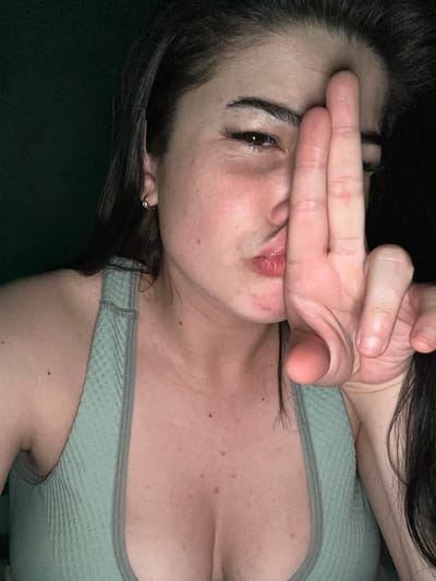 A medium close-up shot shows a young woman with dark hair and fair skin, posing for a selfie in a dimly lit environment. She is wearing a light green textured athletic top. Her right hand is raised to her face, with her index and middle fingers extended and pressed against her forehead and nose, obscuring part of her face. Her lips are pursed, and her left eye is visible, looking towards the camera. The background is dark and out of focus, with a hint of green on the left side. There are no discernible landmarks or details to indicate a specific location, including Gavrilov Posad, Russia. The overall impression is an intimate, casual self-portrait.