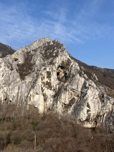 The image captures a large, rugged rock formation or mountain face in the vicinity of Lyutibrod, Bulgaria. The dominant rock is light gray to off-white, characterized by prominent vertical fissures, deep crevices, and darker mineral staining or shadow patterns. Several dark, recessed openings consistent with cave entrances are visible, particularly in the upper-left and central-right portions of the rock face. Sparse, bare deciduous vegetation, including small trees and shrubs, grows on the mountain's ledges and lower slopes. The foreground consists of dense, dormant deciduous brush and leafless trees. A blue sky with scattered wispy white clouds occupies the upper portion of the frame. Multiple thin, dark horizontal lines, identified as overhead cables or wires, traverse the image horizontally across the mid-ground and upper-left, with several small, reddish, rectangular objects resembling cable car gondolas suspended from these lines in the upper-left quadrant. A wooden utility pole is partially visible in the lower-center foreground. No people, animals, or observable actions or interactions are present.