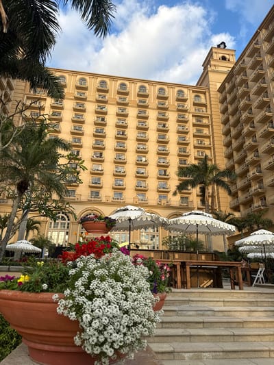 The image displays an exterior view of a multi-story, light-tan architectural complex, identified as a resort or hotel, located in Pelican Bay, USA, under a partly cloudy blue sky. The primary building facade features numerous rows of windows, each equipped with a small wrought-iron balcony. The uppermost floor exhibits arched window openings, also with matching balconies. To the right, a taller building section culminates in a tower flying a black flag with a skull and crossbones (Jolly Roger). Adjacent to this, another building wing with repeating balconies is visible. In the foreground, large terracotta-colored planters are filled with white, red, purple, and yellow flowering plants; white sweet alyssum cascades over the edges of a prominent planter. Wide stone steps descend towards an outdoor patio area. This area is furnished with multiple white, two-tiered umbrellas with scalloped edges, alongside wooden tables and chairs. A white, multi-tiered fountain is partially visible near the base of the building, between the umbrellas. Several tall palm trees and other tropical foliage are interspersed throughout the scene. No individuals are present. The scene is illuminated by natural light, consistent with either morning or late afternoon conditions.