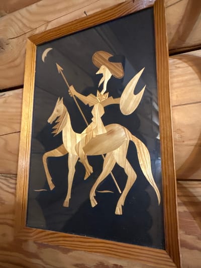 The image is a framed artwork, possibly made of straw or wood shavings, depicting a figure on horseback. The figure wears a wide-brimmed hat and is holding a spear. They are also carrying a shield. A crescent moon is visible in the upper left corner of the artwork. The artwork is mounted on a dark background and framed with a light-colored wooden frame. The background of the image appears to be wooden paneling. The artwork is displayed indoors, on a wall. The location of this artwork in Sergeevka, Russia, is not discernible from the image.