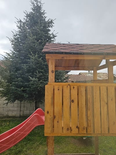 The image displays an outdoor children's play area located in Donchevo, Bulgaria, under an overcast sky. A wooden play structure occupies the right and center of the frame, comprising a raised platform with a vertical wooden plank railing and a gabled roof covered in dark red hexagonal asphalt shingles. A red plastic slide, featuring a subtle wave pattern, extends diagonally downwards from the left side of the play structure to the grassy ground. A grey concrete block wall is visible behind the slide. A large, dense evergreen tree stands prominently in the left background, with distant residential roofs partially visible on the horizon. The ground is covered in green grass. No individuals are present in the scene.