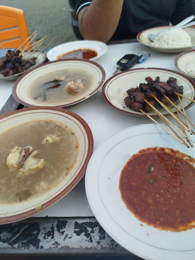 The image captures a close-up, top-down view of a meal laid out on a table, possibly at a casual eatery in Cotpuuk, Indonesia. The table appears to be outdoors, with a hint of a blurry background suggesting an outdoor setting. The foreground is dominated by several plates of food, primarily featuring skewers of grilled meat, likely satay, and bowls of soup.
In the bottom right, a white plate holds a generous portion of a reddish-brown sauce, possibly peanut sauce or a chili-based condiment, with visible texture and small green flecks. To its left, a deeper oval-shaped bowl contains a light-colored broth with chunks of meat, herbs, and what appear to be spices. Further up and to the left, another similar bowl holds a darker, more opaque broth, also with pieces of meat and some visible dark leaves or herbs.
A plate of grilled meat skewers is positioned in the upper middle of the frame. The meat is dark brown and appears well-grilled, with thin wooden skewers protruding from it. Beside this plate, a white bowl is filled with a mound of white rice, and a fork and knife are resting on the rim. Another plate with a smaller portion of sauce is visible further to the right.
A black car key fob with a Toyota logo is placed on the table between the plates of satay and rice, indicating that someone is likely present and may have driven to this location. In the upper left, a portion of an orange plastic chair is visible, suggesting a simple seating arrangement. The table itself has a worn, possibly metal surface with visible peeling paint and dark marks. A person's arm, wearing a black shirt with white trim on the collar, is partially visible in the top background, suggesting their presence at the table. The overall impression is of a hearty, local meal being enjoyed in a relaxed setting.