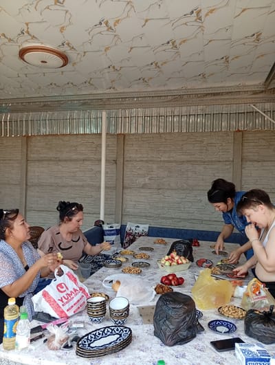 In the city of Teraktagi, Uzbekistan, a group of four women are gathered around a table, sharing a meal outdoors under a patterned ceiling. The setting appears to be a covered patio, with a textured concrete wall serving as the backdrop. The women, dressed casually, are engaged in eating and conversing.

On the table, a spread of food and drinks is laid out. Several plates contain nuts, while a basket holds apples and another dish is filled with what appear to be plums. There are also stacks of decorative bowls and plates, some with intricate blue and white patterns. Two plastic bags, one white with red lettering and another yellow, are visible, along with several water bottles and a bottle of cooking oil. In the foreground, a packet of cigarettes and a mobile phone are also present.

One woman, on the left, is wearing a blue and white patterned top and appears to be eating. Next to her, a woman in a light brown top is also eating. To the right, a woman in a blue dress is reaching for something on the table, while another woman in a white tank top is looking down at the food.

The overall scene suggests a casual gathering, possibly a family meal or a social get-together, taking place in a home's outdoor living space. The environment is bright and seems to be during daylight hours.
