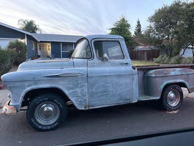 In Roseville, California, an aged, light blue Chevrolet 3100 pickup truck with visible rust and a faded patina is parked on an asphalt road in front of a blue house. The truck's hood is slightly ajar, and the driver's side door is open, revealing a peek of the interior. The truck has chrome hubcaps and Michelin LTX A/T tires. A rose bush with pink and red blooms is situated near the front of the house, and trees and a wooden fence are visible in the background. The sky above is overcast with wispy clouds, suggesting it is either early morning or late evening.