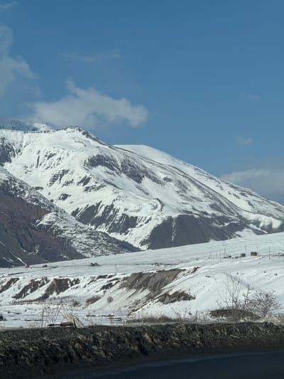 The image depicts a snow-covered mountainous landscape in Kanobi, Georgia, under a partially cloudy blue sky. Snow-covered mountains with exposed rock faces dominate the background. A wide, snow-covered plain with undulating terrain and exposed dark earth occupies the mid-ground. Utility poles and lines traverse this mid-ground. Multiple vehicles are visible on a snow-covered track: two red trucks on the left, and a cluster of lighter-colored vehicles centrally. The immediate foreground consists of a dark asphalt road and a dark, textured embankment. No individuals are discernible.