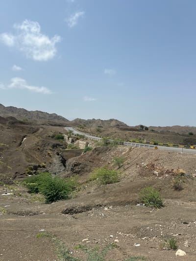 The image shows a desolate, arid landscape in Pakistan with a paved road winding through hills. The terrain is characterized by dry earth, rocks, and sparse vegetation, suggesting a mountainous or hilly region. The road is bordered by a guardrail and appears to be in good condition, with some signage visible. There is evidence of human activity, including some debris scattered along the roadside and in the dry streambeds. The sky is clear blue with a few scattered clouds. The overall impression is one of a remote and rugged environment.