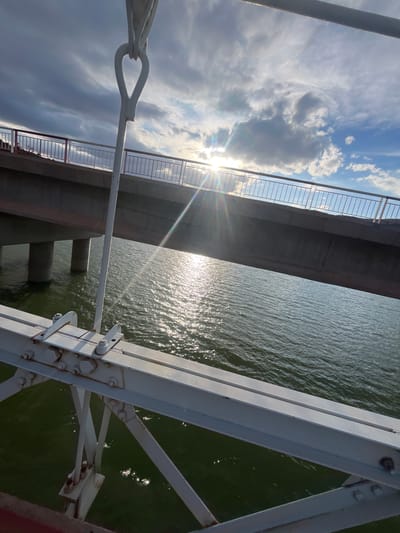The image shows a view from a bridge in Ait Mazigh, Morocco. A section of the bridge railing is visible on the left, with the sun and clouds in the background. Below the bridge is water. The structural supports of the bridge are visible on the right.